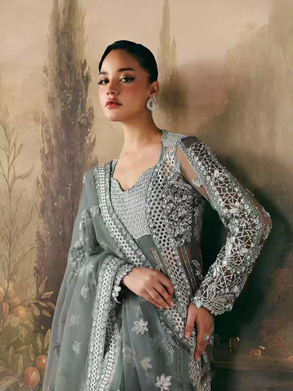 A woman models a traditional South Asian outfit featuring a sage green anarkali-style dress with intricate silver embroidery, a matching embroidered blouse, and a sheer dupatta draped over her shoulder, set against a painterly, warm-toned background.